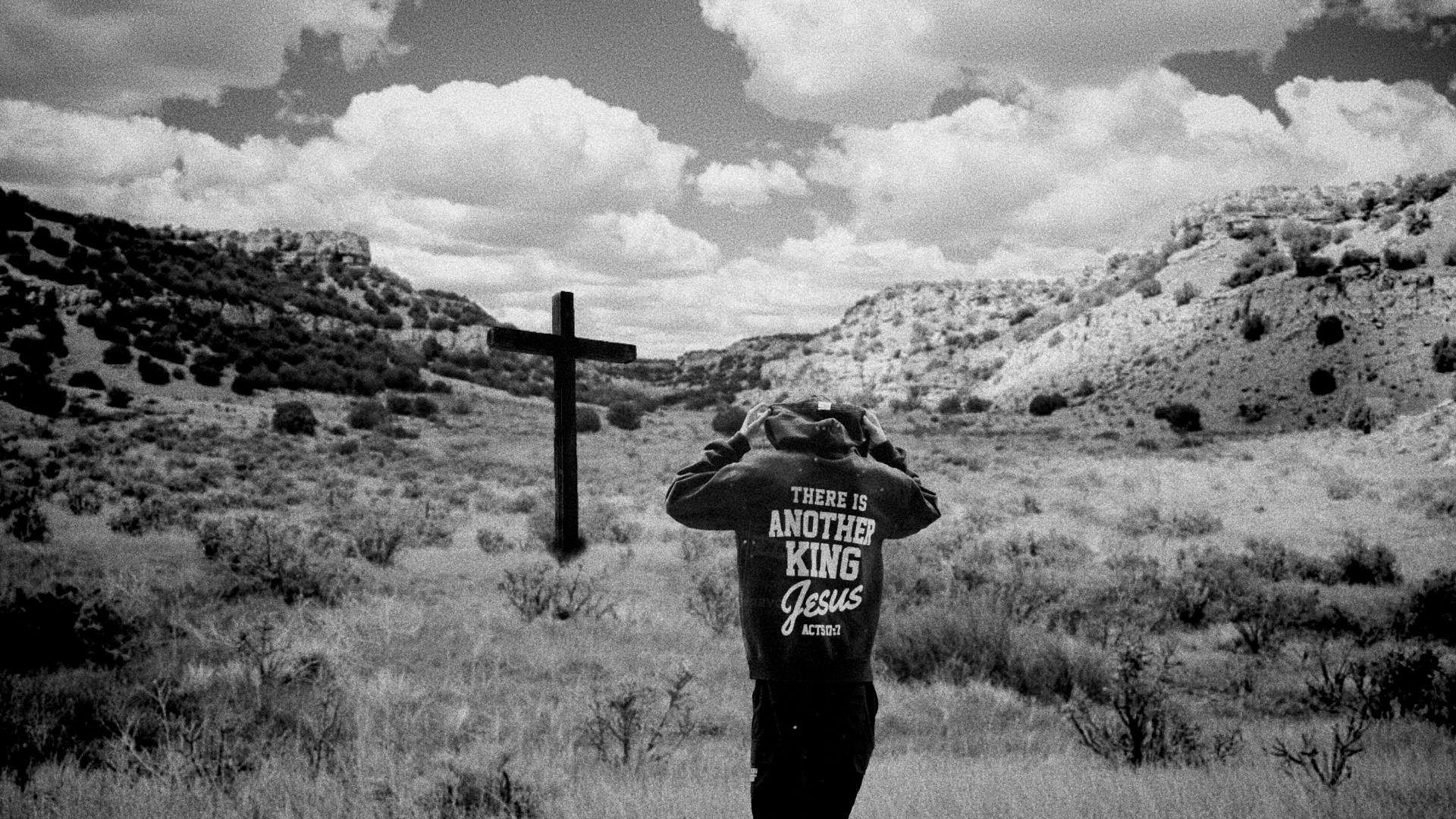 A person stands near a wooden cross in a wide desert valley.